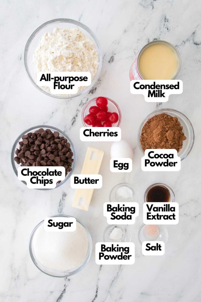 Ingredients for chocolate cherry cookies on a marble countertop: all-purpose flour, condensed milk, cherries, cocoa powder, chocolate chips, butter, egg, sugar, baking soda, vanilla extract, baking powder, salt.