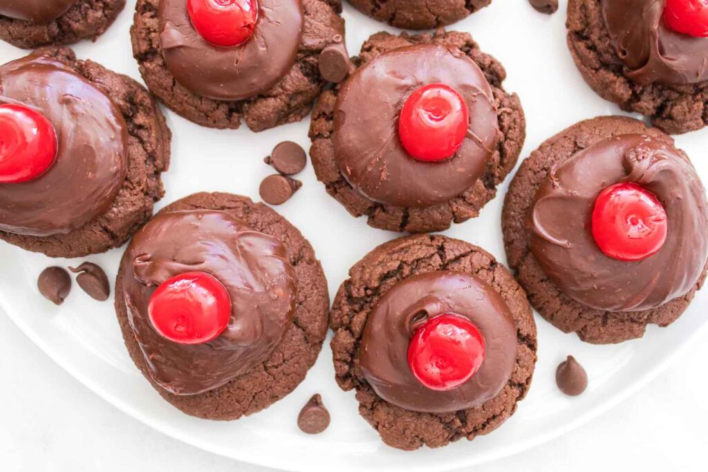 Chocolate cherry cookies topped with rich chocolate icing and adorned with a vibrant red cherry sit elegantly on a pristine white plate.