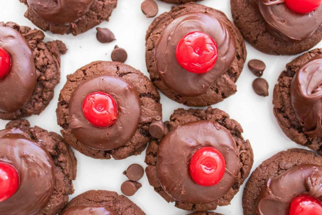 Chocolate cherry cookies topped with rich chocolate frosting and a red cherry, arranged closely together on a white surface with a few scattered chocolate chips.