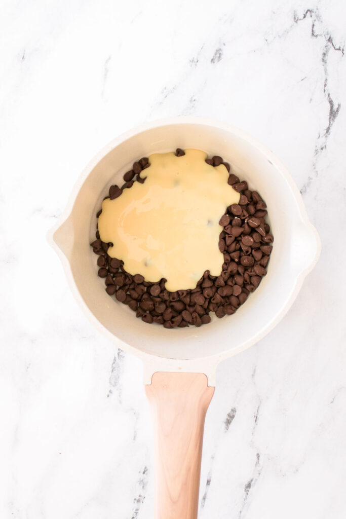 A saucepan sits on a marble surface, filled with chocolate chips melting into a yellow liquid, hinting at the beginnings of rich chocolate cherry cookies.