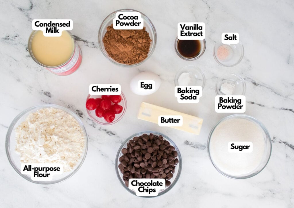 A flat lay of labeled baking ingredients for chocolate cherry cookies, including condensed milk, cocoa powder, vanilla extract, salt, baking soda, baking powder, sugar, chocolate chips, cherries, butter, egg, and flour.