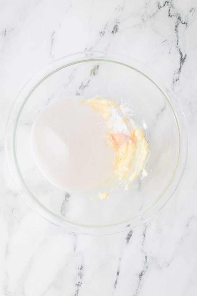 A clear glass bowl contains butter, sugar, eggs, flour, and a hint of chocolate on a marble countertop, ready to become delicious cherry cookies.