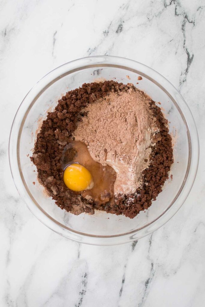 A glass bowl with brownie mix, cocoa powder, an egg, and the enticing aroma of chocolate cherry cookies on a marble surface.