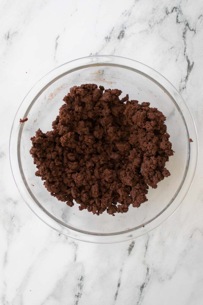 A glass bowl containing crumbly, chocolate cherry cookie dough rests on a white marble surface.