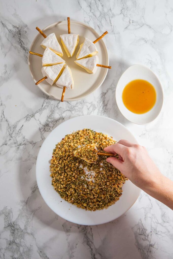 A hand dips a wedge of brie with pretzel sticks into a nut mixture, crafting festive Christmas cheese trees. Nearby, a bowl of honey and a plate with cheese slices on pretzel sticks rest elegantly on the marble surface.