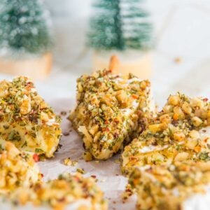 Close-up of brie cheese covered with a festive mixture of nuts and herbs, arranged on parchment paper with small decorative Christmas trees in the background.