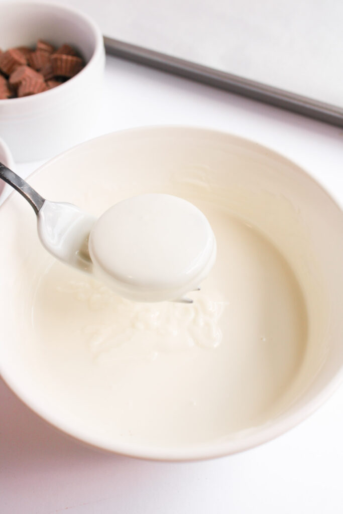 A spoon holds a round Oreo cookie dipped in a bowl of white chocolate, transforming it into a festive Christmas treat. A small bowl of chocolate pieces is in the background, adding to the holiday charm of these ornament-inspired delights.