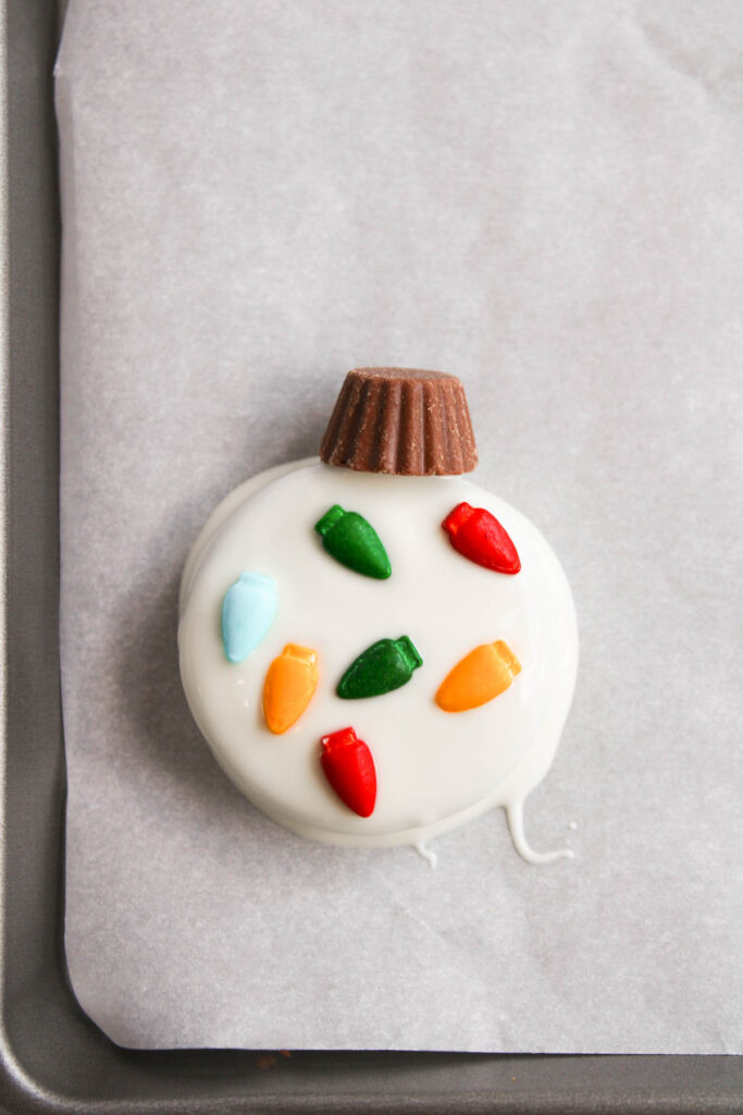 A round cookie coated with white icing, adorned with colorful candy pieces and topped with a peanut butter cup resembling a festive Oreo Christmas ornament, all placed delicately on parchment paper.