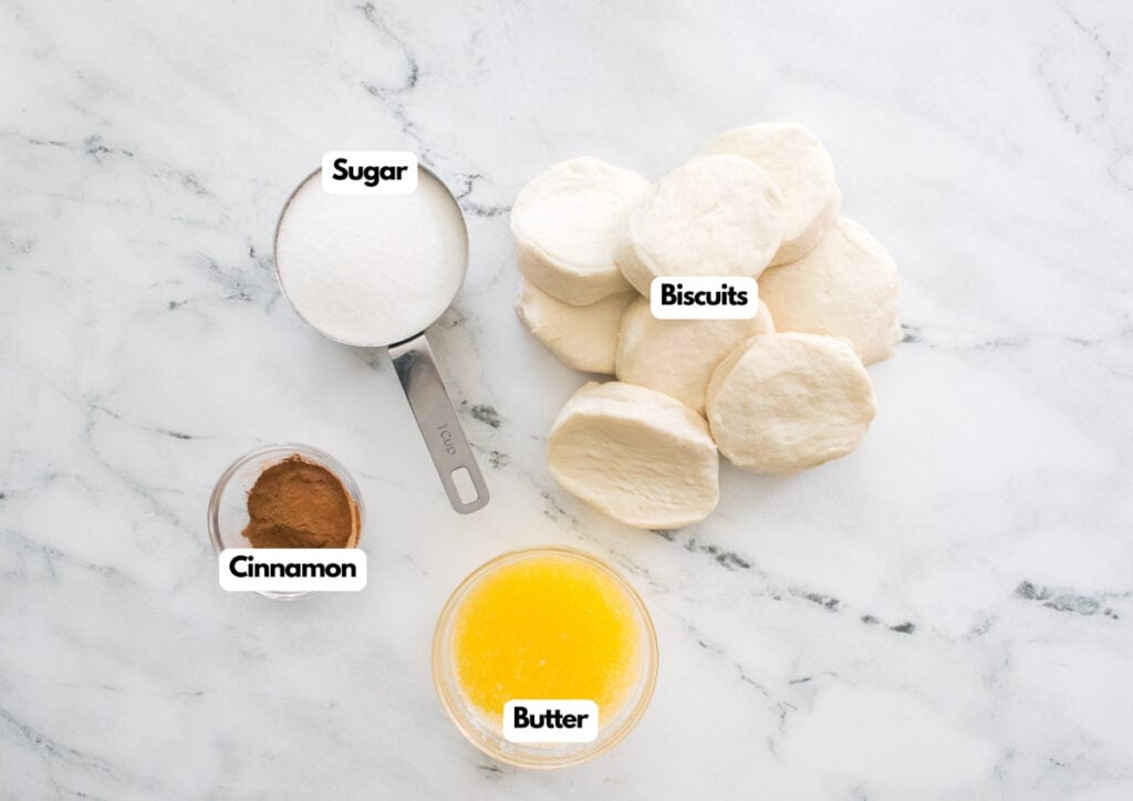 Overhead view of measured ingredients for Cinnamon Sugar Biscuit Bites on a marble surface: sugar, cinnamon, melted butter in a bowl, and several uncooked biscuits.