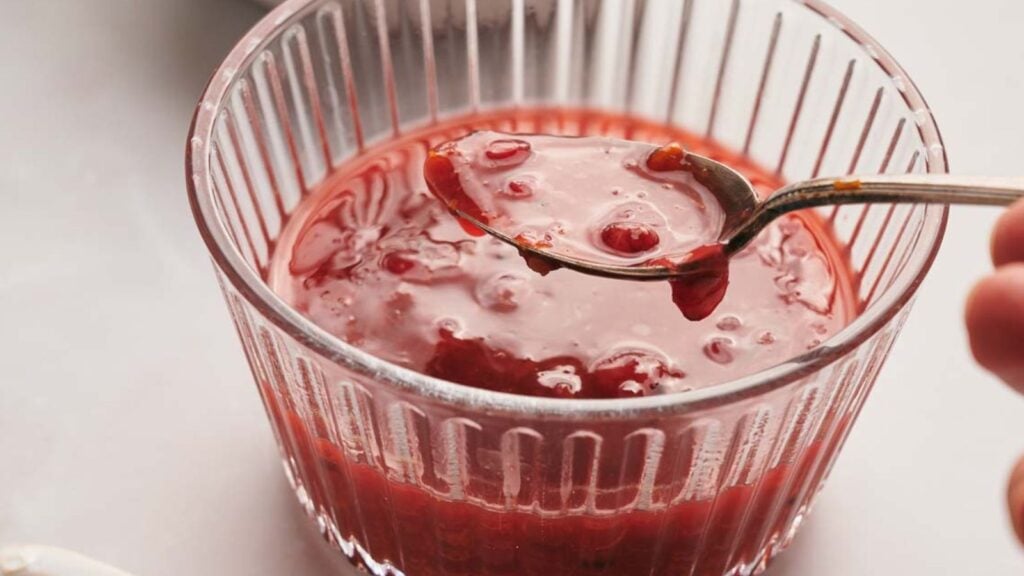 A hand holds a spoon scooping cranberry sauce from a clear glass bowl.