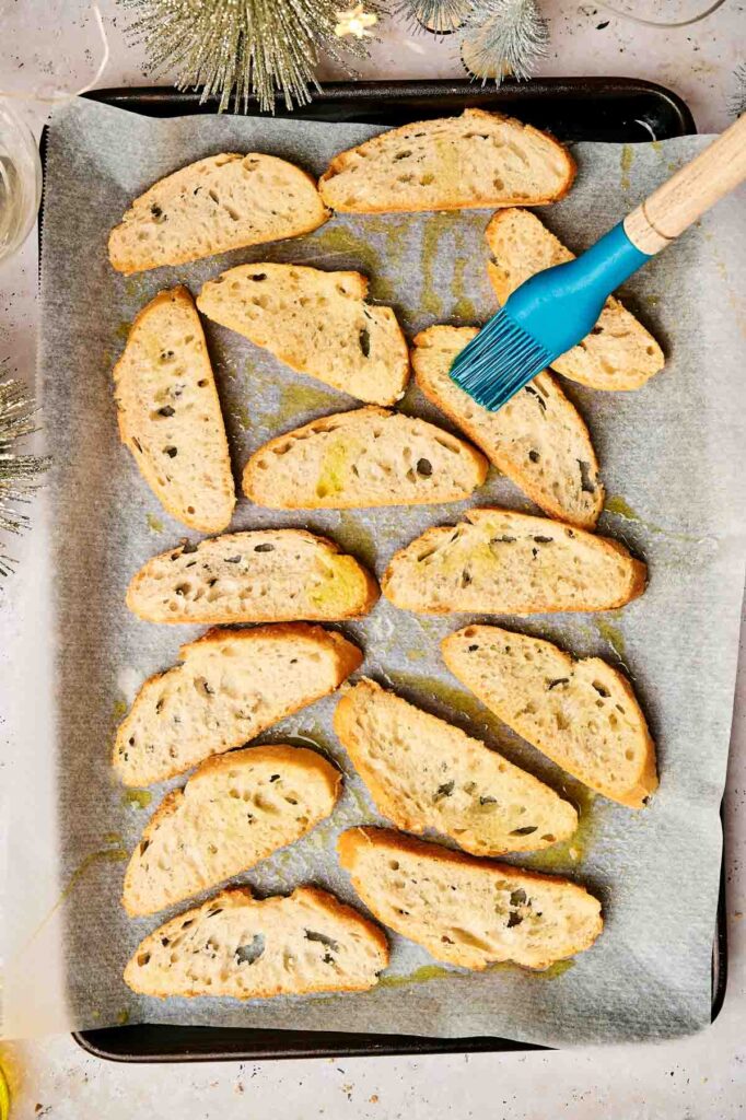 A baking tray lined with parchment paper holds slices of bread being brushed with olive oil, perfect for making party crostini. A blue pastry brush is visible on the right.
