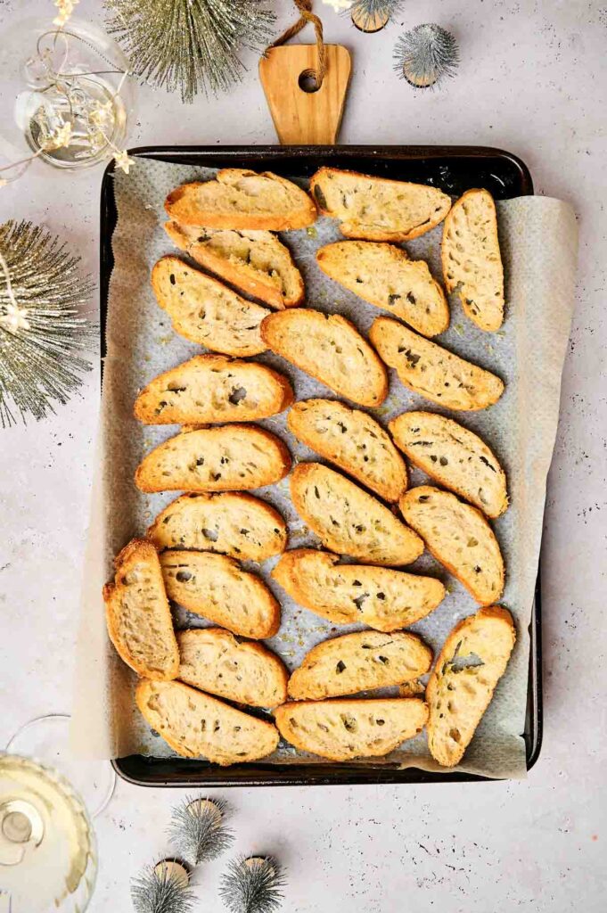 A tray of party crostini slices rests on parchment paper, surrounded by small Christmas decorations and two glasses of white wine.