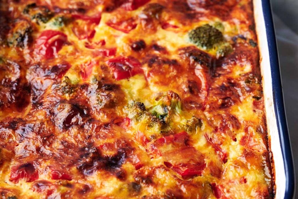 Close-up of a baked vegetable and chicken noodle casserole with a golden, slightly charred cheese topping, showing pieces of broccoli and tomatoes.