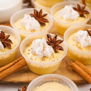 Small cups of creamy eggnog pudding shots topped with whipped cream, star anise, and cinnamon powder are arranged on a wooden tray with cinnamon sticks around.