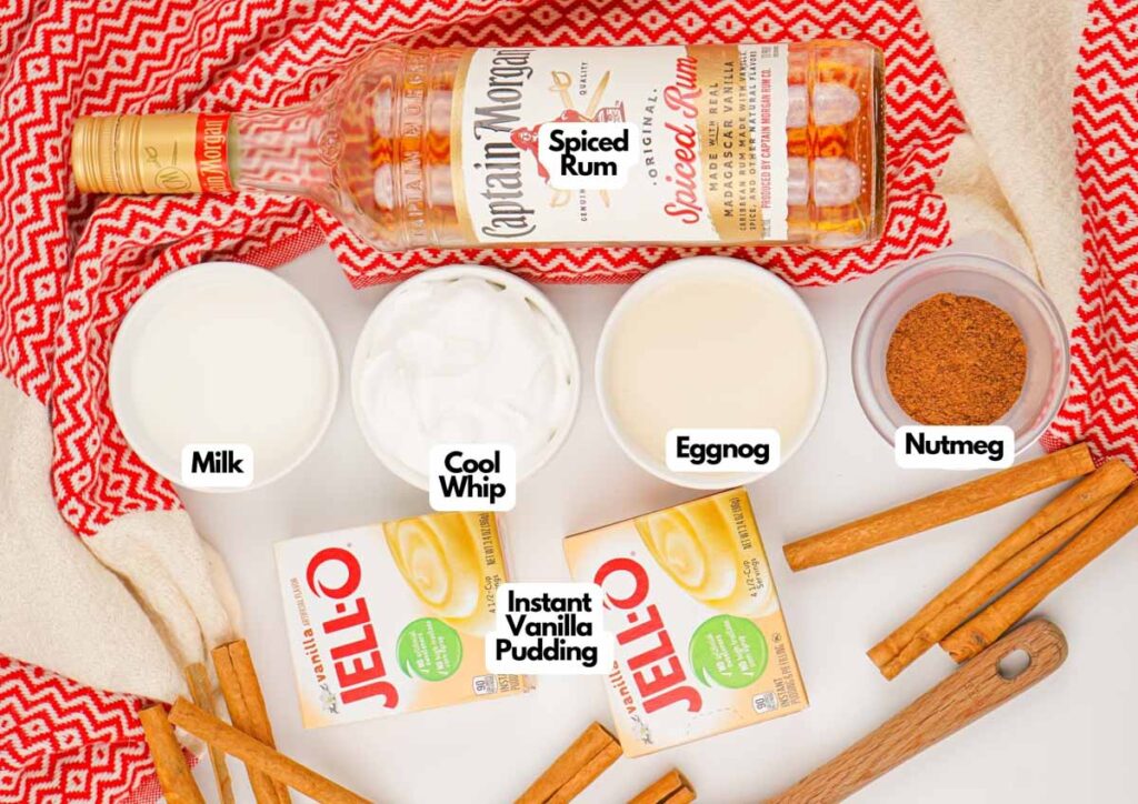 Ingredients for Eggnog Pudding Shots laid out on a table: spiced rum, milk, Cool Whip, eggnog, nutmeg, cinnamon sticks, and instant vanilla pudding mix.