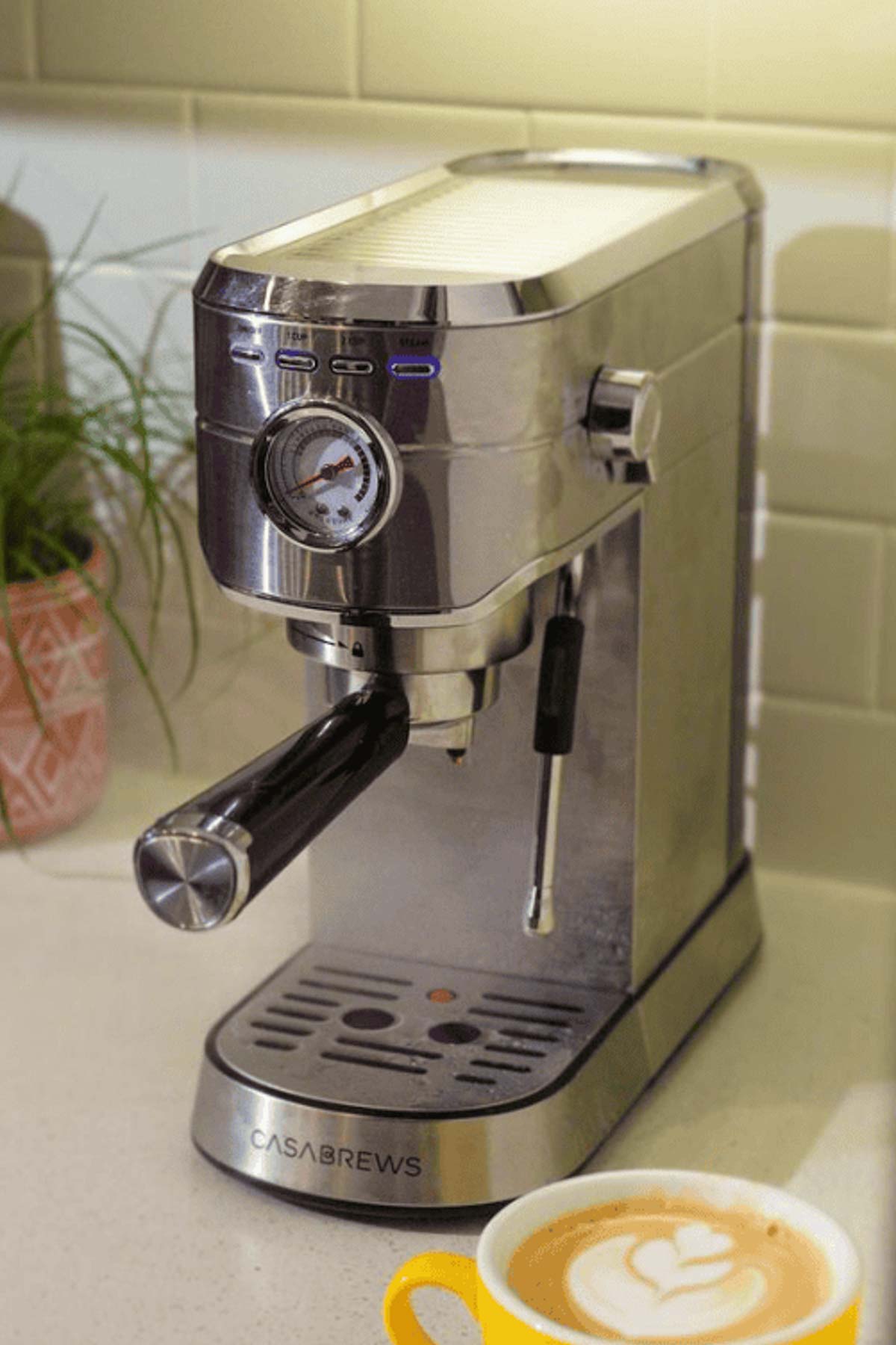 A stainless steel espresso machine with a steam wand and pressure gauge sits on a countertop next to a cappuccino with latte art.