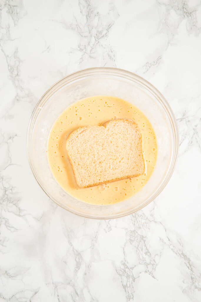 A slice of bread soaking in a bowl of beaten eggs on a marble countertop.
