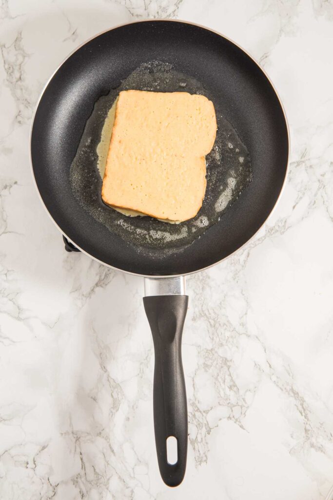 A slice of bread being toasted in a black frying pan on a marble countertop.