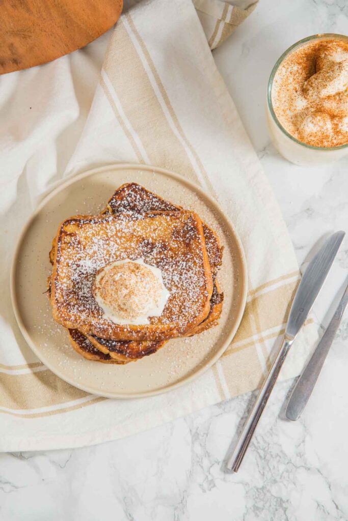 A plate of French toast topped with powdered sugar and cream, next to a drink with whipped cream and a sprinkle of cinnamon, set on a marble surface with a fork and knife.