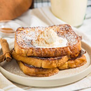 Three slices of French toast with whipped cream and powdered sugar on a plate, next to cinnamon sticks. A mug of a creamy beverage is blurred in the background.
