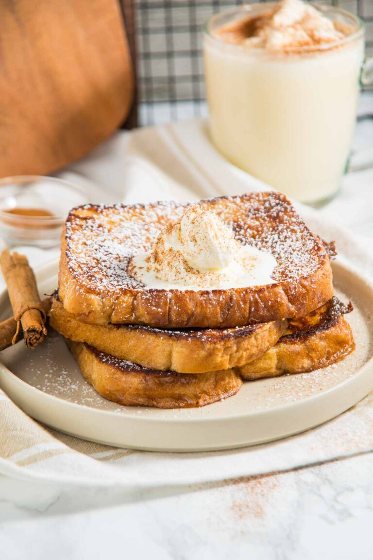 Three slices of French toast with whipped cream and powdered sugar on a plate, next to cinnamon sticks. A mug of a creamy beverage is blurred in the background.