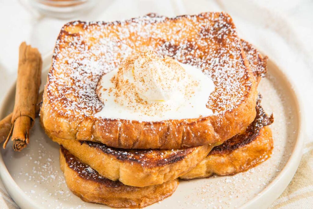 A stack of French toast topped with whipped cream and powdered sugar on a plate, with cinnamon sticks beside it.