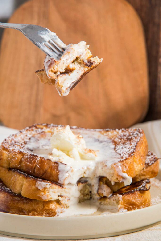 Fork holding a bite above a stack of syrup-drizzled French toast topped with melting butter and powdered sugar on a plate.
