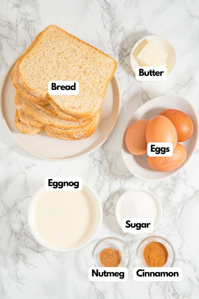 Ingredients on a marble surface: slices of bread, butter, eggs, a bowl of eggnog, sugar, nutmeg, and cinnamon.