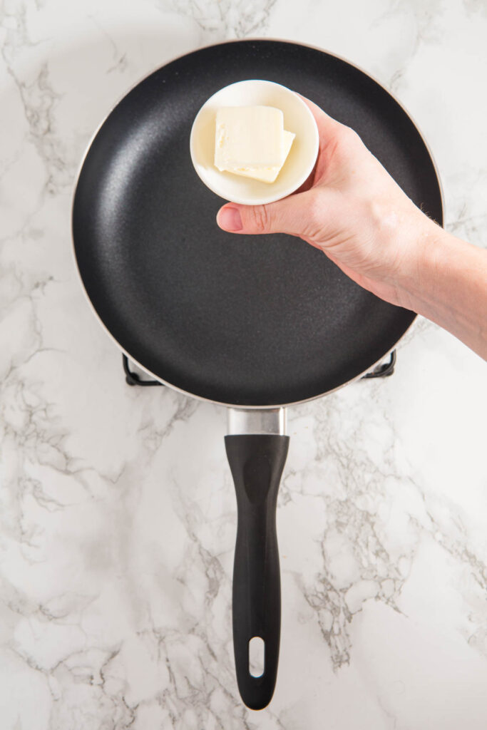 A hand holding a small white bowl with butter above a black frying pan on a marble countertop.