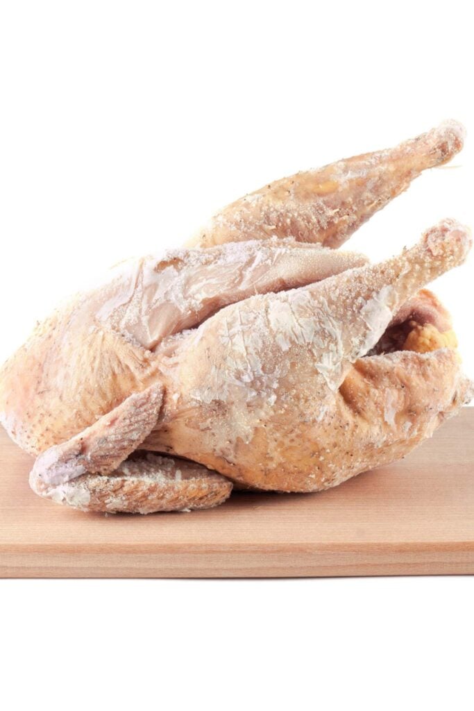 A frozen whole chicken rests on a wooden cutting board, reminding one of how to thaw a turkey with care and patience.