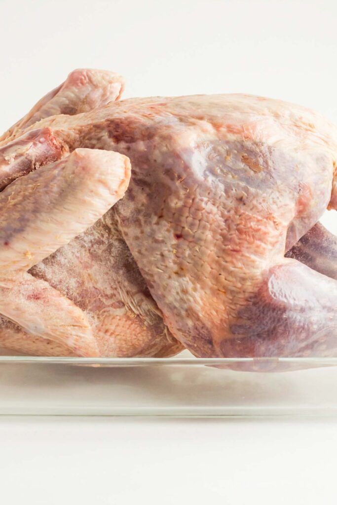 A raw whole turkey rests in a glass dish on a white background, waiting to be transformed into a delicious feast. Understand how to thaw a turkey properly to ensure your meal is both safe and succulent.