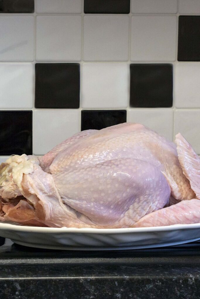 A raw whole chicken is placed on a white platter, set against a black and white tiled background. This inviting scene might remind one of learning how to thaw a turkey for a perfect holiday feast.