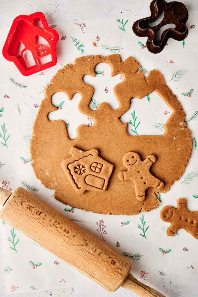Gingerbread dough rests on a festive paper with cutout shapes of a gingerbread man and house, perfect for making air fryer gingerbread cookies. Red and brown cookie cutters and a rolling pin are nearby, ready for baking magic.