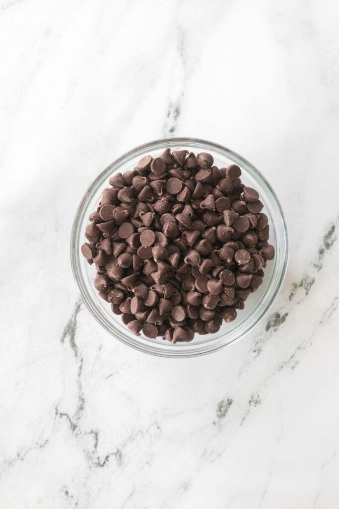 A glass bowl filled with dark chocolate chips, perfect for crafting homemade buckeyes, rests elegantly on a marble surface.