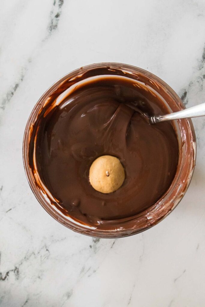A homemade buckeye cookie being dipped in a bowl of melted chocolate with a spoon resting on the cool marble surface.
