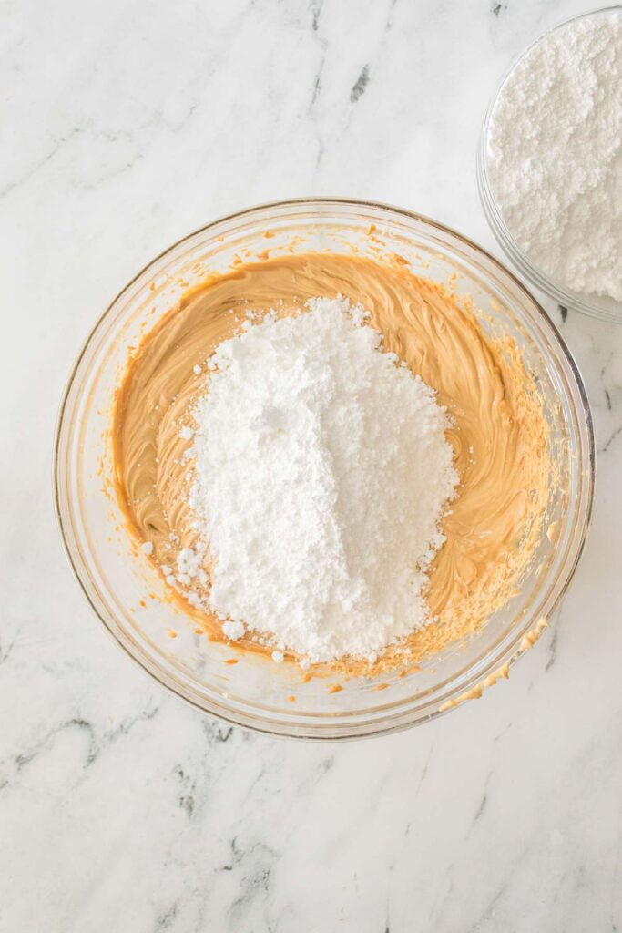 A glass bowl contains creamy peanut butter mixed with a mound of powdered sugar, reminiscent of homemade buckeyes. In the background, another bowl brims with more powdered sugar.