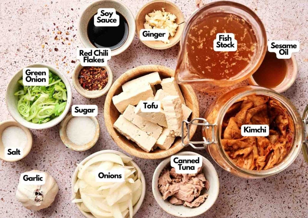 A variety of labeled ingredients for Kimchi Stew, including tofu, kimchi, fish stock, canned tuna, green onion, onions, garlic, salt, sugar, soy sauce, red chili flakes, ginger, and sesame oil.