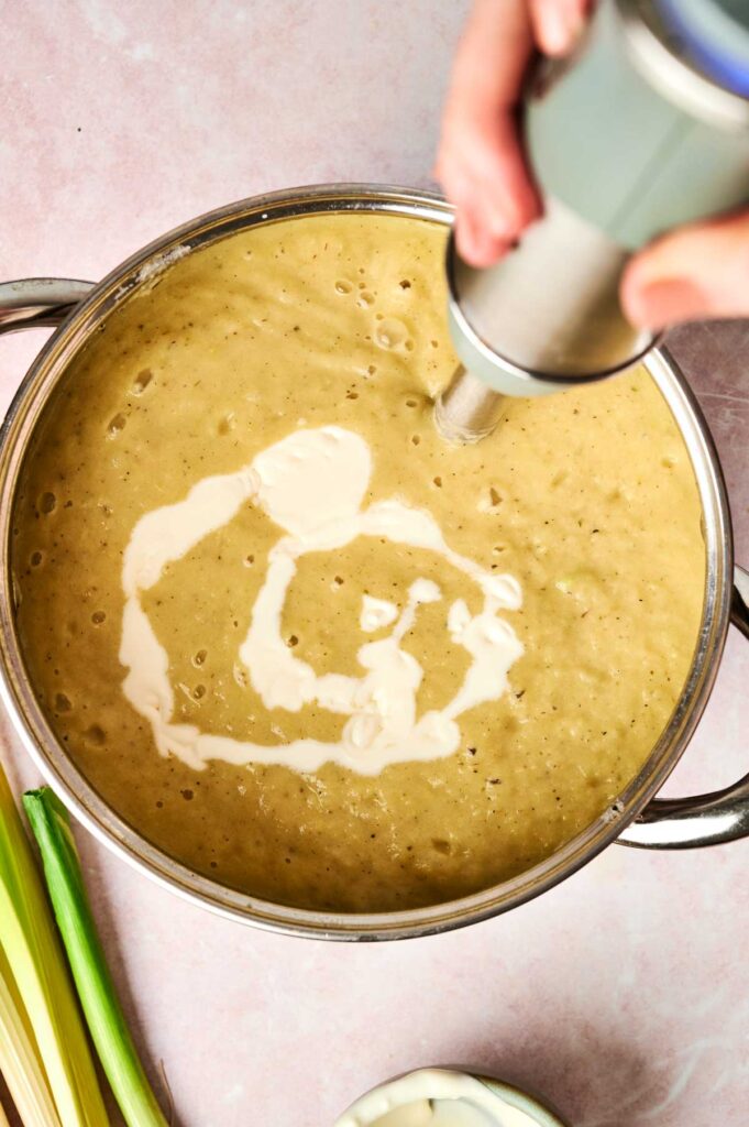 A person blending creamy leek and potato soup in a pot with an immersion blender, adding cream for richness. Nearby, stalks of celery await their turn.