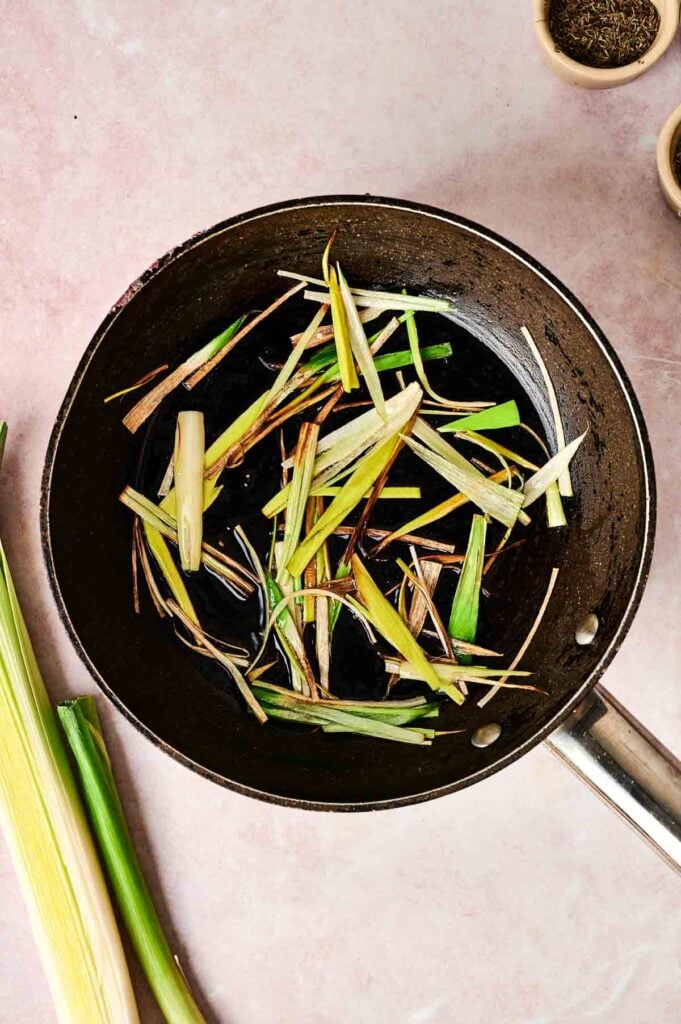 Pan with lemongrass in oil on a light surface, evoking the comforting aroma of leek and potato soup, with more lemongrass and small containers in the background.