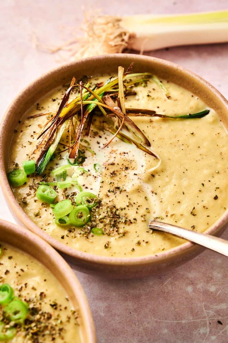 Bowl of creamy leek and potato soup topped with sliced green onions, herbs, and crispy julienned vegetables. A spoon rests in the bowl.