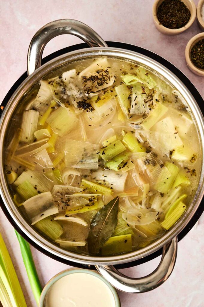 A pot of leek and potato soup simmers gently, with chopped leeks, creamy potatoes, herbs, and a hint of bay leaf adding to the inviting aroma.