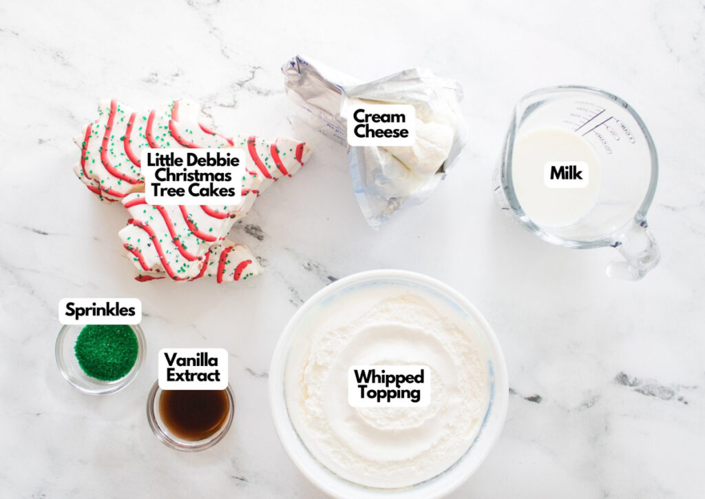 Ingredients on a marble surface for little debbie christmas tree dip: Christmas tree cakes, cream cheese, milk in a measuring cup, green sprinkles, vanilla extract, and a bowl of whipped topping.