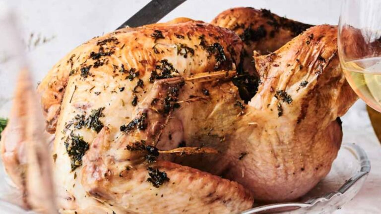 Roasted turkey with herbs on a glass platter, partially carved, surrounded by glasses with a light beverage.