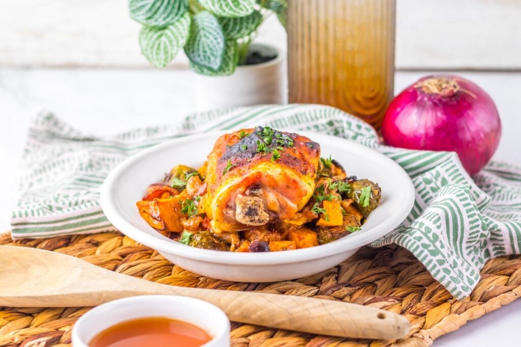 A bowl of Maple Dijon Chicken Sheet Pan is served over mixed vegetables, placed on a woven mat with a wooden spoon, red onion, sauce, and a potted plant in the background.