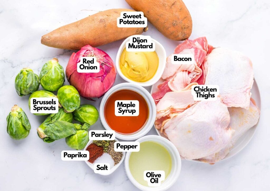 Ingredients on a countertop for Maple Dijon Chicken Sheet Pan include sweet potatoes, brussels sprouts, red onion, parsley, paprika, salt, pepper, Dijon mustard, maple syrup, olive oil, bacon, and chicken thighs.