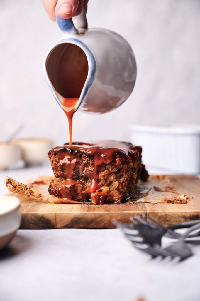 A person pours sauce over a square piece of bread pudding on a wooden board.
