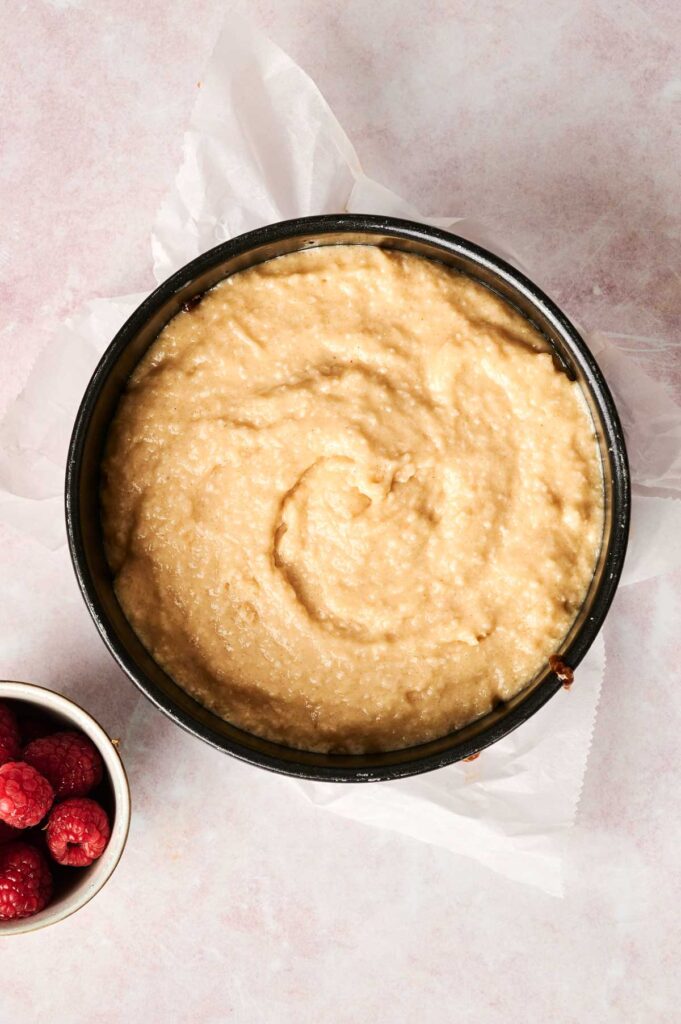 A round cake pan filled with unbaked batter on a parchment-lined surface, next to a small bowl of raspberries.
