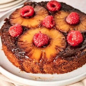 A pineapple upside-down cake topped with pineapple slices and raspberries on a light-colored plate. Plates and spoons are in the background.