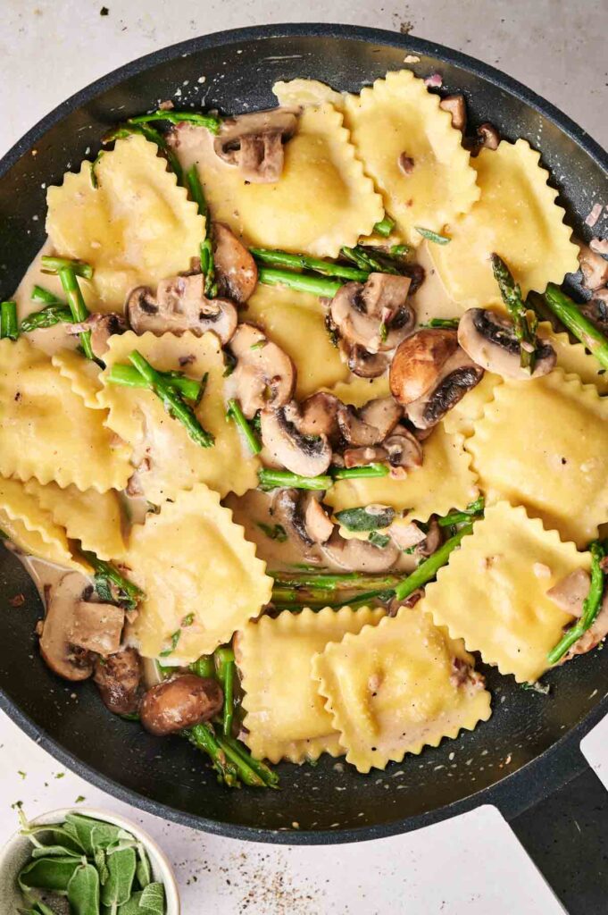 Ravioli casserole with mushrooms and asparagus in a creamy sauce, all nestled in a black pan.
