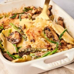 A ravioli casserole with mushrooms, green beans, and a golden cheese crust, all nestled in a rectangular white baking dish with a serving spoon.